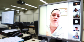 Mulher fala, em destaque, em reunião remota, na tela de um computador. Do lado direito da tela estão seis outras janelas menores, com imagens de dois homens, duas mulheres e duassomente com iniciais de nomes.