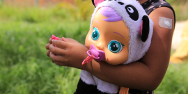 Criança ao ar livre segura uma boneca nas mãos. A boneca, de grandes olhos azuis, usa uma chupeta rosa e uma touca de bichinho