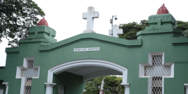 Entrada em arco do Cemitério do Bonfim. A entrada é pintada de verde e tem duas cruzes vazadas na edificação com detalhes em branco. Ao fundo, é possível avistar os túmulos