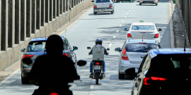 Duas motos e três carros trafegam em viaduto, durante o dia. Uma das motos está entre dois carros. 