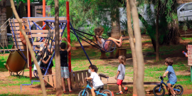 Seis crianças brincam em brinquedos ao ar livre, cercadas de verde e acompanhadas por um adulto.