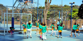 Dez crianças treinam basquete acompanhadas de dois adultos, durante o dia, em local aberto. 