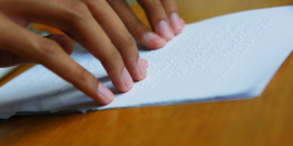 As mãos de uma pessoa percorrem um papel branco escrito em braile