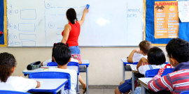 Professora, de costas, escreve em quadro branco; atrás delas, seis crianças, sentadas em carteiras, a observam.