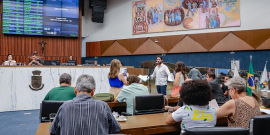 A foto é plano aberto mostra o Plenário Amintas de Barros. Visto de trás, em primeiro plano, está um grupo de vereadores sentados. O vereador Gabriel Azevedo está em pé, virado de frente, falando ao microfone, ele usa blusa social branca e calça jeans. Mais atrás, em um nível acima, estão sentados o presidente da reunião e a secretária, e na parede acima deles o telão que contabiliza a presença e os votos.