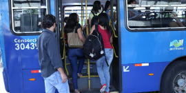 A foto mostra a lateral da parte de traseira de um ônibus azul. A porta está aberta, com pessoas dentro e outras preste a entrar.