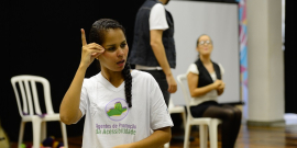 Mulher com camisa branca escrito "Agentes de promoção da acessbilidade", faz um sinal em Libras 