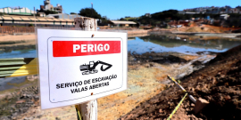 Foto de barragem ao fundo com placa a frente escrito "serviço de escavação valas abertas"