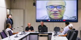 Foto de reunião, com convidados sentados à mesa e uma pessoa no telão na parede atrás.