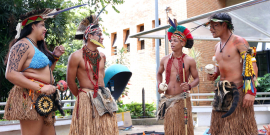 Pessoas indígenas tocam instrumentos de percussão em vestes tradicionais na frente da entrada da CMBH