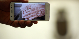 Imagem na tela de um celular de mãos caucasianas, com marcas do tempo, postas sobre o colo  
