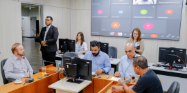 Vereadores estão sentados no plenário Camil Caram. O telão atrás deles mostra a videochamada com um parlamentar presente virtualmente.