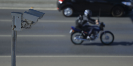 Foto mostra, em primeiro plano, câmera de monitoramento e, na rua, moto e automóvel
