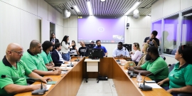 Vereadores e convidados da audiência estão sentados no plenário. O telão atrás deles mostra a videochamada com participantes presentes virtualmente.
