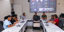 Vereadores e convidados da audiência estão sentados no plenário. O telão atrás deles mostra a videochamada com participantes presentes virtualmente.