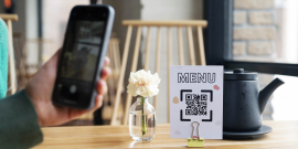 Mão com celular fazendo leitura de QR code em cima de uma mesa