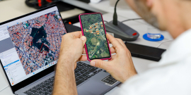 homem com o celular na mãe e o computador à frente, ambos com imagem geográfica de satélite