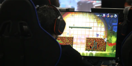Na foto, no canto esquerdo, observa-se parcialmente a parte de trás de uma cadeira e um jovem sentado utilizando um fone de ouvido. Na sua frente a tela de um computador mostra um jogo.