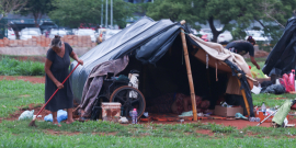 Mulher em situação de rua varre terreno ao lado de cabana improvisada com lona