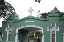 Entrada em arco do Cemitério do Bonfim. A entrada é pintada de verde e tem duas cruzes vazadas na edificação com detalhes em branco. Ao fundo, é possível avistar os túmulos