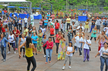 Mais de 40 pessoas praticam atividade física em quadra de escola, orientados por uma mulher, durante o dia. 