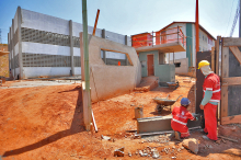 Dois homens de capacete e uniforme laranja da Prefeitura de Belo Horizone, trabalham em obra, erguendo parede