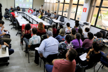 Mais de vinte pessoas sentadas acompanham reunião com oito homens e mulheres sentados à mesa.