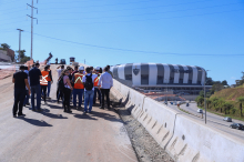 Parlamentares e equipes sobem uma via asfaltada e ainda em fase de construção, construída como contrapartida, que dá acesso à Arena. Ao fundo é possível ver a cúpula do Estádio