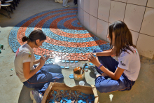 Duas garotas, sentadas, completam mosaico de cerâmica no chão à mão, durante o dia
