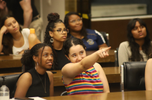 Seis jovens sentadas, uma delas aponta para frente e conversa com a garota ao seu lado