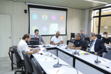 Cinco homens sentados à mesa, em reunião presencial.