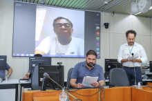 Parlamentar, sentado na sala, em reunião virtual com outro parlamentar, exibido em telão.
