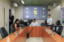 Dois parlamentares sentados à mesa, em reunião presencial, acompanhados por parlamentares que participam remotamente, exibidos em telão, ao fundo.