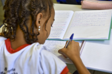 Criança negra de uniforme, de costas sentada na carteira, escreve em caderno com caneta azul.