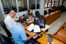 Vereadores discutem projetos no plenário. O computador na frente deles mostra videochamada com vereadores presentes virtualmente.