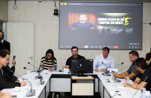 Vereadores e convidados assentados no Plenário Helvécio Arantes durante audiência pública. O telão atrás deles mostra apresentação.