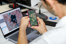homem com o celular na mãe e o computador à frente, ambos com imagem geográfica de satélite